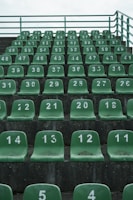 Rows of green stadium seats are arranged in ascending order. Each seat is numbered in white, and the numbers appear to be worn. The seating is on a grey concrete structure, and a metal railing is visible at the top.