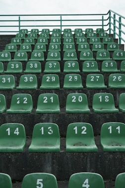 Rows of green stadium seats are arranged in ascending order. Each seat is numbered in white, and the numbers appear to be worn. The seating is on a grey concrete structure, and a metal railing is visible at the top.