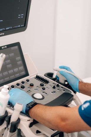 A person wearing blue gloves is operating an ultrasound machine. The control panel is visible with various buttons and dials, and two screens display information. There is also a small bottle in a holder next to the machine.