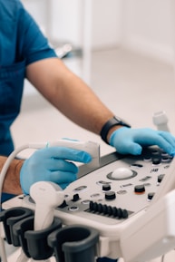 A person wearing blue medical scrubs and gloves operates an ultrasound machine in a clinical setting. The individual's hands are adjusting the controls and handling the transducer probe, which is resting on the console equipped with various buttons and dials.