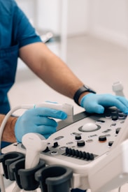 A person wearing blue medical scrubs and gloves operates an ultrasound machine in a clinical setting. The individual's hands are adjusting the controls and handling the transducer probe, which is resting on the console equipped with various buttons and dials.