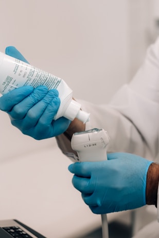 A person wearing blue gloves is applying a gel from a bottle onto a medical device, possibly an ultrasound probe, indicating preparation for a medical procedure.
