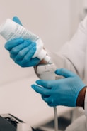 A person wearing blue gloves is holding a white container of gel and applying it to a medical ultrasound device. The individual is dressed in a white lab coat, indicating a professional or clinical setting.