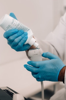 A person wearing blue gloves is holding a white container of gel and applying it to a medical ultrasound device. The individual is dressed in a white lab coat, indicating a professional or clinical setting.