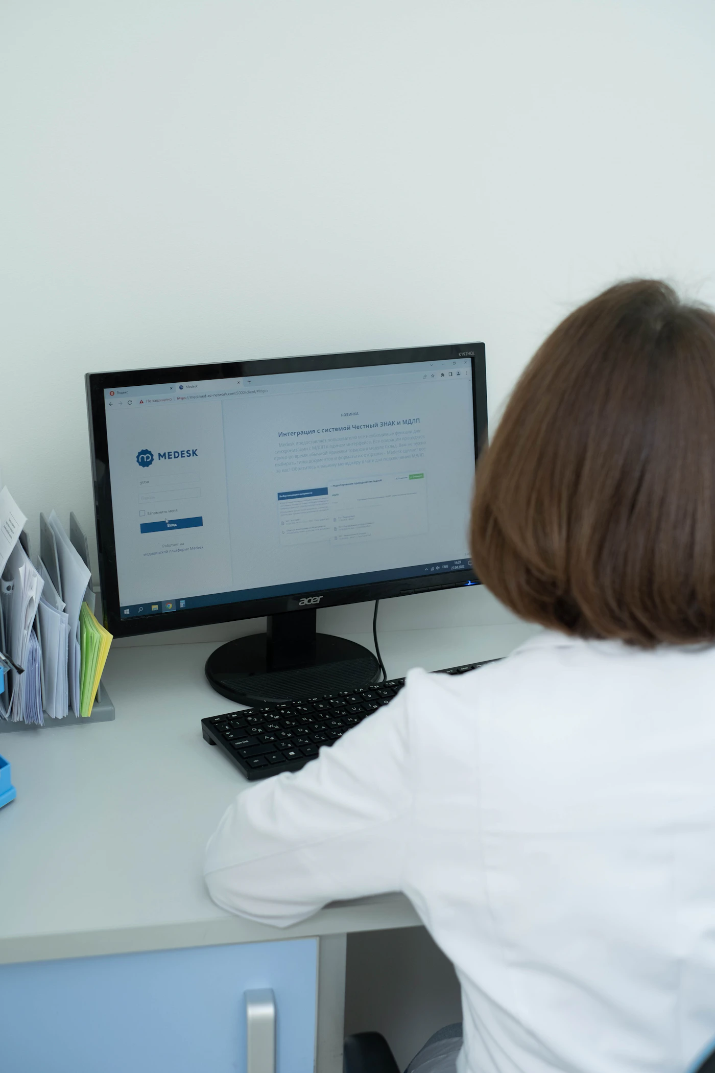 A clinician in a white coat reviewing a patient record on a secure screen at a tidy office desk, with paper files organized beside them