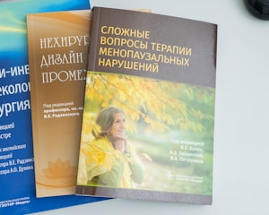 Three books are placed on a flat surface. The book on the left has a blue cover with text in a foreign language. The middle book has a brown cover and also features text along with an image of a lotus flower. The book on the right has a cover featuring an older woman in a thoughtful pose, with autumn leaves in the background and text in a foreign language.