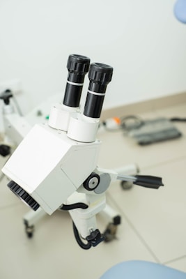 A white microscope with two eyepieces is placed on a light-colored tiled floor. The microscope is equipped with black dials and has an intricate mechanical design. Cables and connectors are visible, suggesting it's part of a larger setup. The background contains blurred elements, including a wall and other indistinct equipment.