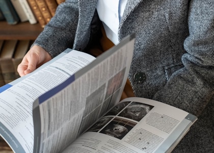 A person wearing a grey herringbone patterned coat is holding and reading an open magazine or book. The pages contain text and several images, possibly images related to medical or scientific topics. In the background, books are visible on a shelf.