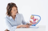 A person wearing a light blue shirt is seated at a white surface, attentively examining a colorful anatomical model of the human digestive system. The model is detailed, showing various organs in vibrant pink, yellow, and blue colors.