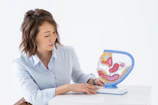 A person wearing a light blue shirt is seated at a white surface, attentively examining a colorful anatomical model of the human digestive system. The model is detailed, showing various organs in vibrant pink, yellow, and blue colors.