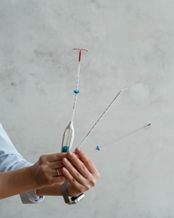 A hand holding three medical instruments with long, thin handles and small attachments at the end. One of the instruments has a red plastic part forming a T-shape, another has a blue ring and a white stick, and the third appears to have a clear loop at the end. The background is a plain, light-colored wall.