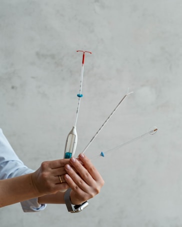 A hand holding three medical instruments with long, thin handles and small attachments at the end. One of the instruments has a red plastic part forming a T-shape, another has a blue ring and a white stick, and the third appears to have a clear loop at the end. The background is a plain, light-colored wall.