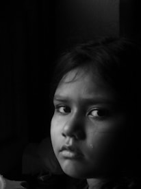 A young child with a solemn expression, looking towards the camera. There is a tear on their cheek, indicating sadness or distress. The image is in black and white, adding a dramatic and emotional effect.