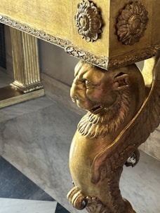 A detailed, ornate piece of furniture featuring a carved lion's head and floral motifs. The surface is gilt, with intricate patterns and designs. The leg of the furniture shows craftsmanship with lion features and decorative elements.