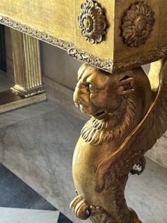 A detailed, ornate piece of furniture featuring a carved lion's head and floral motifs. The surface is gilt, with intricate patterns and designs. The leg of the furniture shows craftsmanship with lion features and decorative elements.