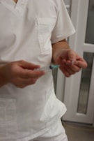 A person dressed in white medical scrubs is holding a syringe with one hand and a small object in the other hand. The background features a blurred interior with white and glass elements, possibly a medical facility.