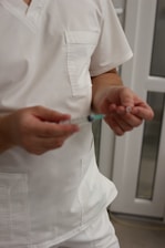 A person dressed in white medical scrubs is holding a syringe with one hand and a small object in the other hand. The background features a blurred interior with white and glass elements, possibly a medical facility.