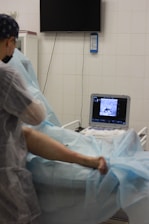 A medical professional is performing a procedure on a patient's leg in a clinical setting. The patient is lying on a table with blue surgical drapes covering most of their body. An ultrasound machine displays an image, possibly of the patient's leg, while a monitor hangs on the wall behind them.