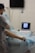 A medical professional is performing a procedure on a patient's leg in a clinical setting. The patient is lying on a table with blue surgical drapes covering most of their body. An ultrasound machine displays an image, possibly of the patient's leg, while a monitor hangs on the wall behind them.