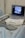 An ultrasound machine displaying an image on its screen is situated on a hospital trolley. The machine has a covered keyboard and several wires or cables attached. The setting appears to be a medical environment, likely a hospital or clinic, with a sterile and clinical appearance.
