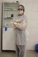 A person wearing a hairnet, face mask, and protective gown stands with arms crossed in a clinical or laboratory setting. Behind them, a cabinet contains various medical supplies.