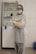 A person wearing a hairnet, face mask, and protective gown stands with arms crossed in a clinical or laboratory setting. Behind them, a cabinet contains various medical supplies.
