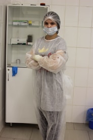 A person wearing a hairnet, face mask, and protective gown stands with arms crossed in a clinical or laboratory setting. Behind them, a cabinet contains various medical supplies.
