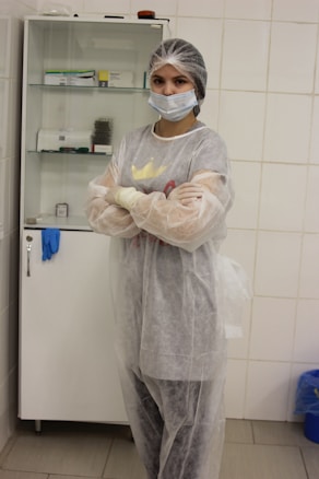 A person wearing a hairnet, face mask, and protective gown stands with arms crossed in a clinical or laboratory setting. Behind them, a cabinet contains various medical supplies.
