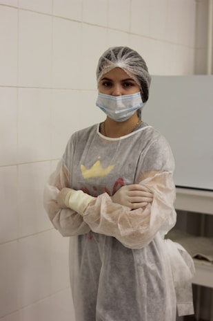 A person in a laboratory environment wearing a disposable gown, face mask, and hair cover. The individual stands with arms crossed in a tiled room.