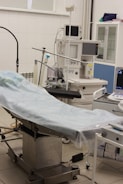 The scene depicts an operating room or medical setup with a surgical table covered in a light blue sterile sheet. Surrounding the table are various pieces of medical equipment, including monitoring devices and anesthesia machines. Shelves in the background hold additional medical supplies.