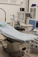 The scene depicts an operating room or medical setup with a surgical table covered in a light blue sterile sheet. Surrounding the table are various pieces of medical equipment, including monitoring devices and anesthesia machines. Shelves in the background hold additional medical supplies.
