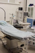 The scene depicts an operating room or medical setup with a surgical table covered in a light blue sterile sheet. Surrounding the table are various pieces of medical equipment, including monitoring devices and anesthesia machines. Shelves in the background hold additional medical supplies.