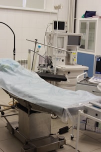 The scene depicts an operating room or medical setup with a surgical table covered in a light blue sterile sheet. Surrounding the table are various pieces of medical equipment, including monitoring devices and anesthesia machines. Shelves in the background hold additional medical supplies.
