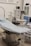 The scene depicts an operating room or medical setup with a surgical table covered in a light blue sterile sheet. Surrounding the table are various pieces of medical equipment, including monitoring devices and anesthesia machines. Shelves in the background hold additional medical supplies.