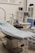The scene depicts an operating room or medical setup with a surgical table covered in a light blue sterile sheet. Surrounding the table are various pieces of medical equipment, including monitoring devices and anesthesia machines. Shelves in the background hold additional medical supplies.