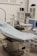 The scene depicts an operating room or medical setup with a surgical table covered in a light blue sterile sheet. Surrounding the table are various pieces of medical equipment, including monitoring devices and anesthesia machines. Shelves in the background hold additional medical supplies.