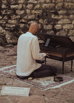 A person is kneeling on a patterned rug while playing a clavicytherium, an ancient keyboard instrument, in front of a rustic stone wall. The individual is wearing a white shirt and dark pants, and a small sign is placed on the ground providing information about the instrument.