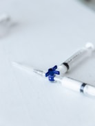 Two syringes lie on a flat white surface, arranged with their needles pointing outward. The syringes have a clear body and one features a blue rotary valve in the center.