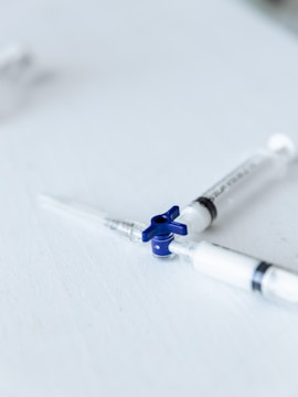 Two syringes lie on a flat white surface, arranged with their needles pointing outward. The syringes have a clear body and one features a blue rotary valve in the center.