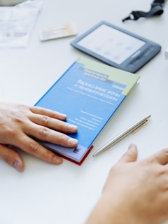 A person's hands are resting on a table beside a blue book with Cyrillic text on the cover. Next to the book is a silver pen, an e-reader, and a blurred card, all placed on a light surface. The scene suggests a workspace or study area.