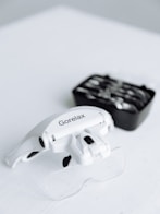 A close-up of a white headband magnifier with the brand name 'Gorelax' printed on it, resting on a white surface. In the background, there is a black box containing various lens options that are slightly out of focus.