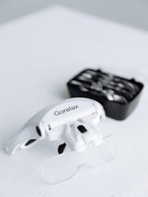 A close-up of a white headband magnifier with the brand name 'Gorelax' printed on it, resting on a white surface. In the background, there is a black box containing various lens options that are slightly out of focus.