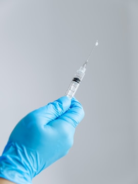 A hand wearing a blue medical glove holding a syringe with a clear liquid. The background is plain and light-colored, drawing focus to the syringe and the glove.