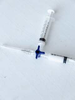 Two medical syringes are placed on a light gray surface, arranged diagonally with one having a blue plunger cap. The focus is sharp, emphasizing the markings and details on the syringes.