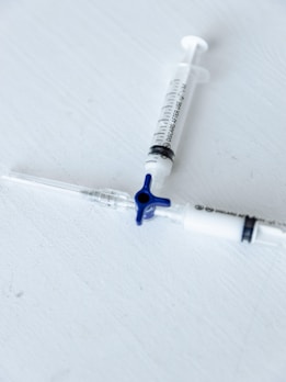 Two medical syringes are placed on a light gray surface, arranged diagonally with one having a blue plunger cap. The focus is sharp, emphasizing the markings and details on the syringes.