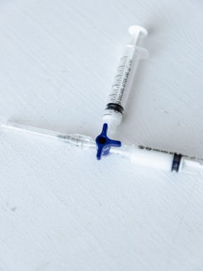 Two medical syringes are placed on a light gray surface, arranged diagonally with one having a blue plunger cap. The focus is sharp, emphasizing the markings and details on the syringes.