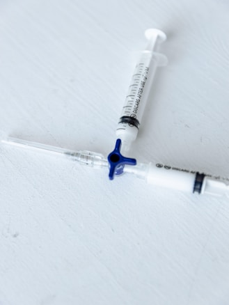 Two medical syringes are placed on a light gray surface, arranged diagonally with one having a blue plunger cap. The focus is sharp, emphasizing the markings and details on the syringes.