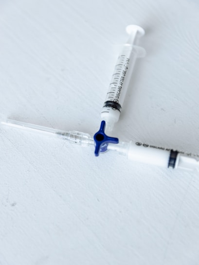 Two medical syringes are placed on a light gray surface, arranged diagonally with one having a blue plunger cap. The focus is sharp, emphasizing the markings and details on the syringes.