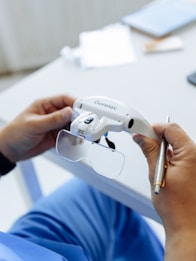 A person is holding a white device labeled 'Gorelax' with attached lenses, which appears to be some form of visual equipment or augmented glasses. The person is also holding a pen. The background includes a blurred desk surface with scattered items.