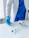 A medical professional wearing a white coat and blue scrubs is handling a scalpel. The individual is also wearing blue gloves. On the table in front are medical instruments, including syringes and a device possibly used for magnification purposes, placed on a clean white surface.